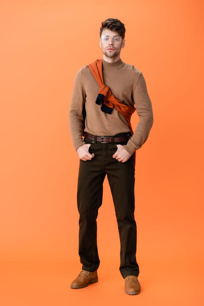 volledige lengte van stijlvolle man in bril staande met handen in zakken op oranje  - Foto, afbeelding