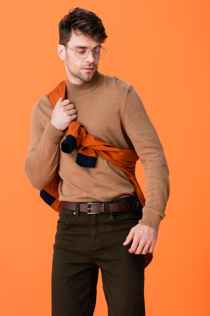 stijlvolle man in bril aanraken jumper en weg te kijken geïsoleerd op oranje  - Foto, afbeelding