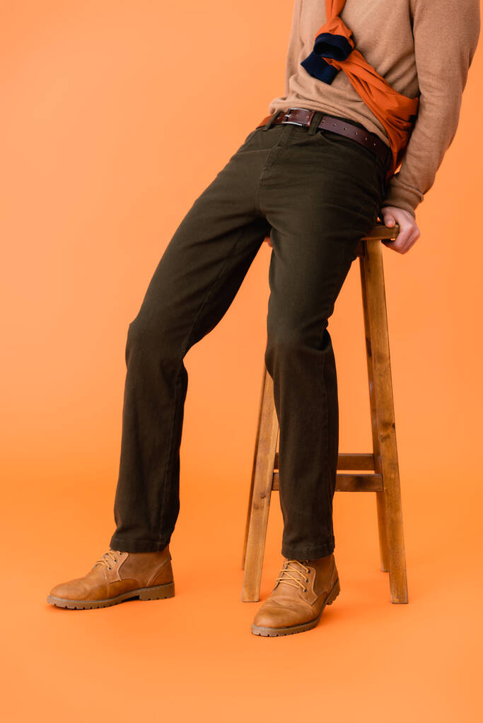 vista ritagliata dell'uomo di tendenza in abito autunnale seduto su sgabello in legno su arancione  - Foto, immagini