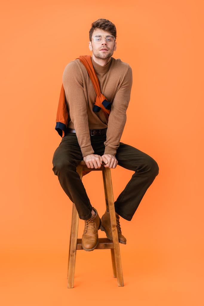 homme à la mode en tenue d'automne et lunettes assises sur tabouret en bois sur orange  - Photo, image