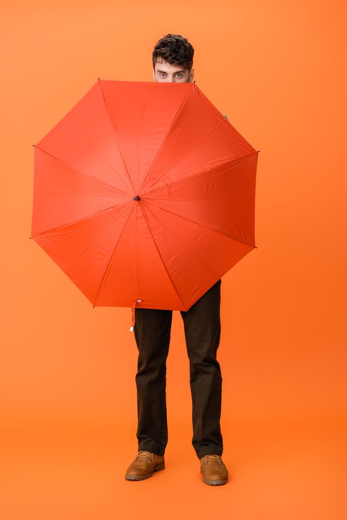 Мужчина в коричневых штанах в полный рост, держащий пуховик на оранжевом  - Фото, изображение