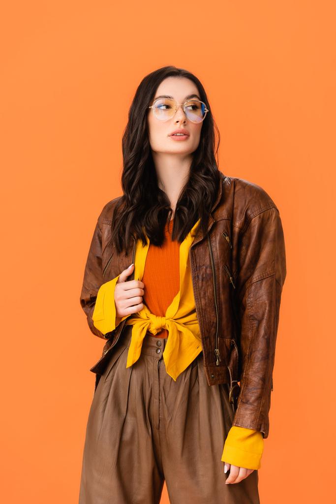 mujer con estilo en traje de otoño y gafas tocando chaqueta y mirando hacia otro lado aislado en naranja  - Foto, Imagen