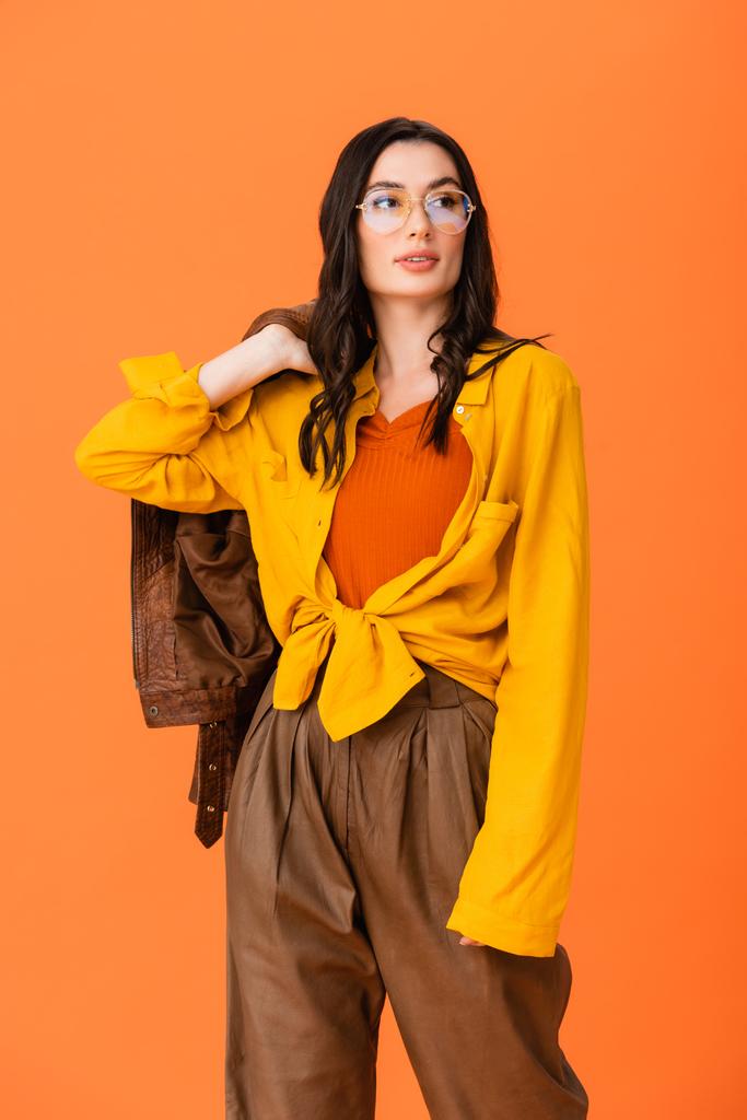 módní žena v brýlích a podzimní oblečení drží koženou bundu izolované na oranžové  - Fotografie, Obrázek