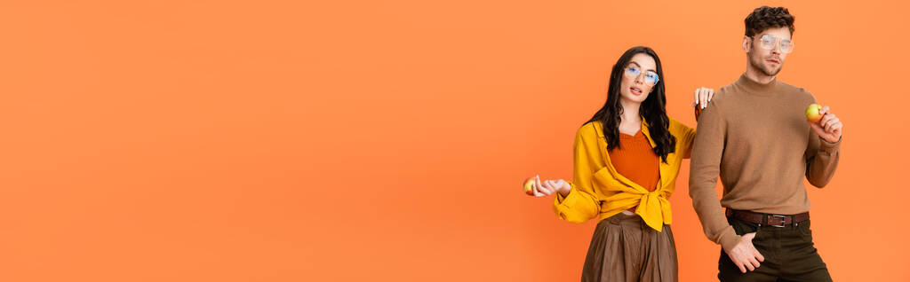 cultivo panorámico de pareja de moda en gafas y traje de otoño con manzanas aisladas en naranja - Foto, Imagen