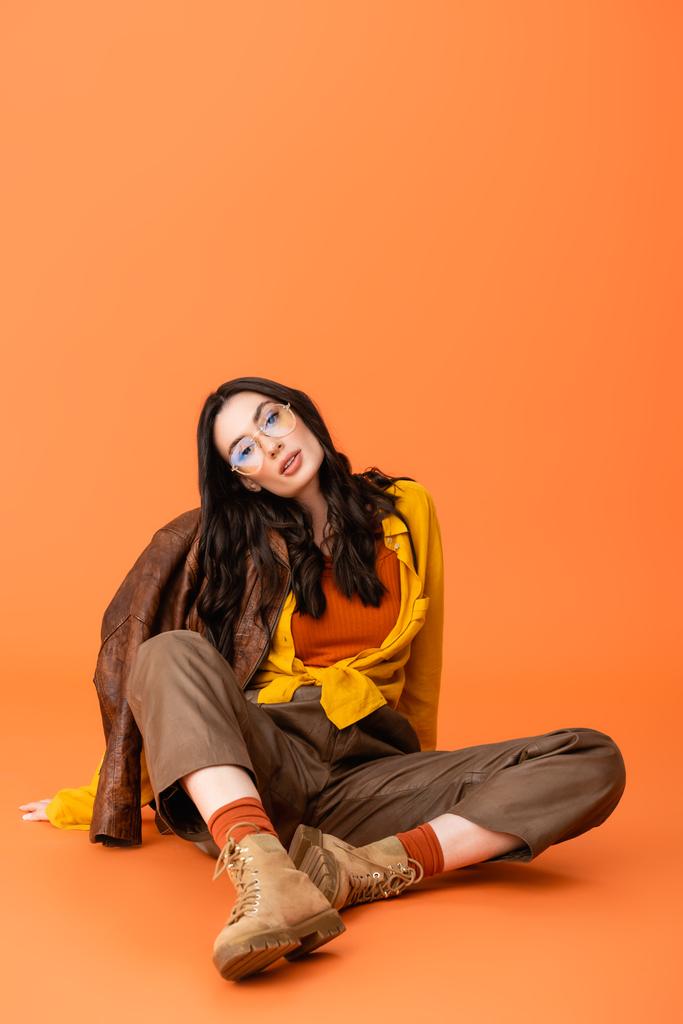 donna di tendenza in occhiali e abbigliamento autunnale e giacca di pelle seduta su arancione  - Foto, immagini