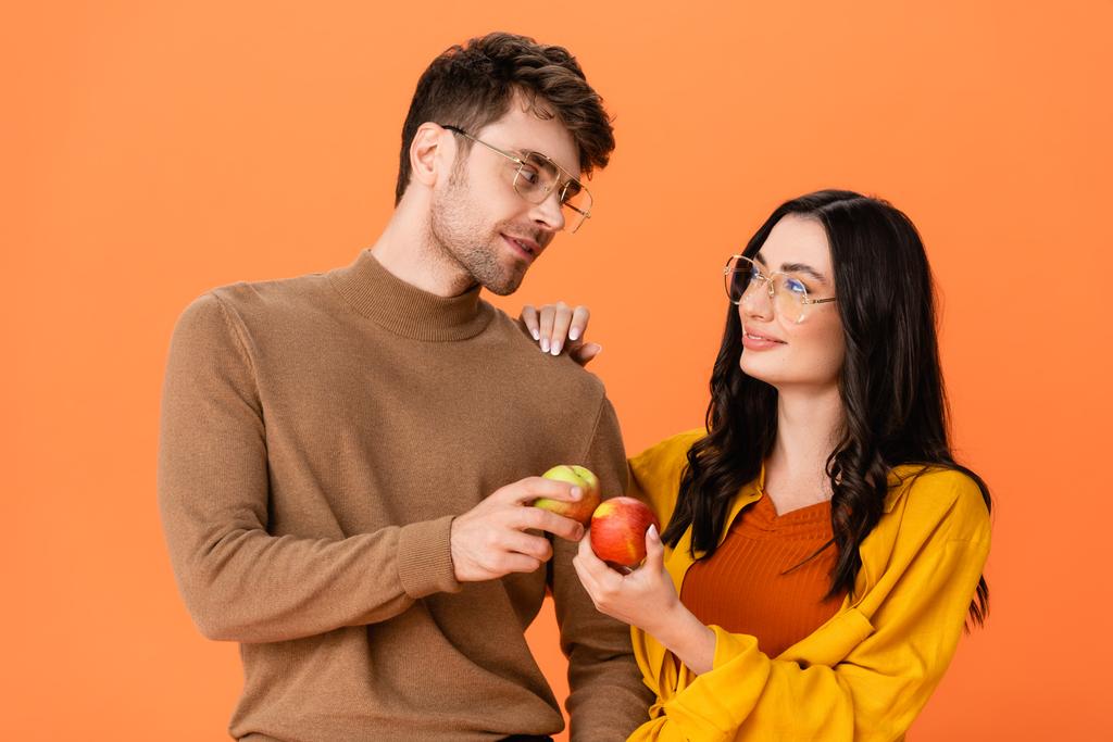 pareja de moda en gafas y traje de otoño sosteniendo manzanas mientras se miran aislados en naranja - Foto, Imagen