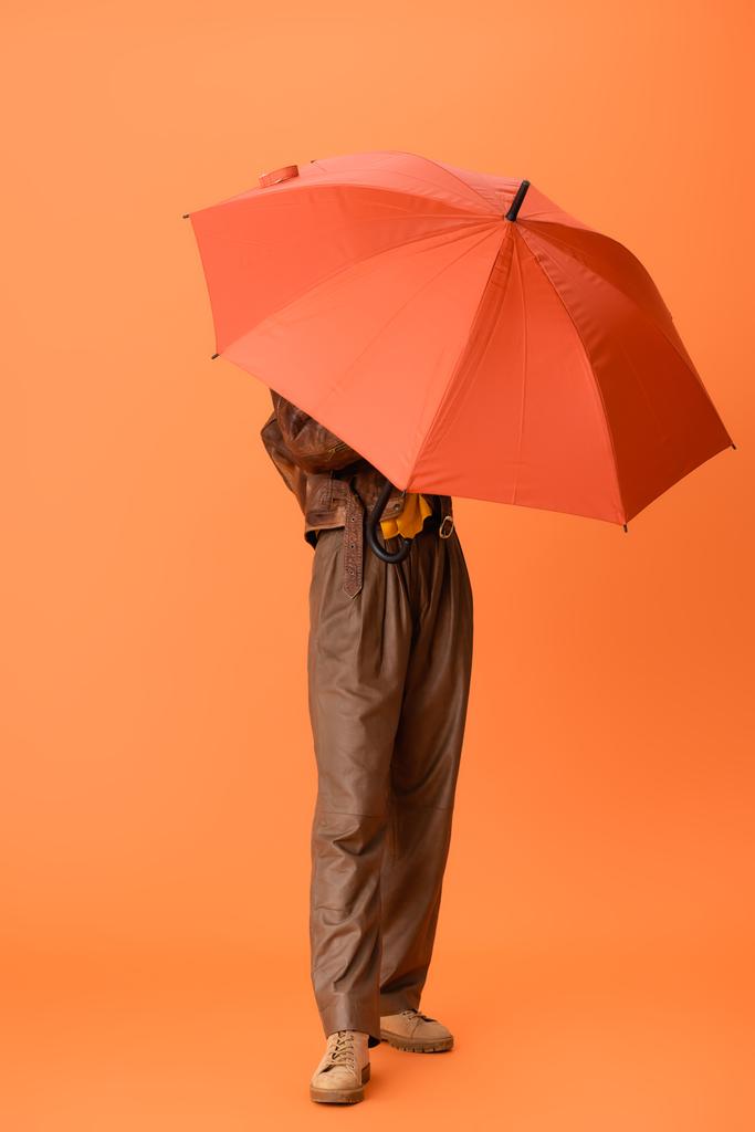 volledige lengte van modieuze vrouw in herfst outfit en laarzen bedekking gezicht met paraplu op oranje - Foto, afbeelding