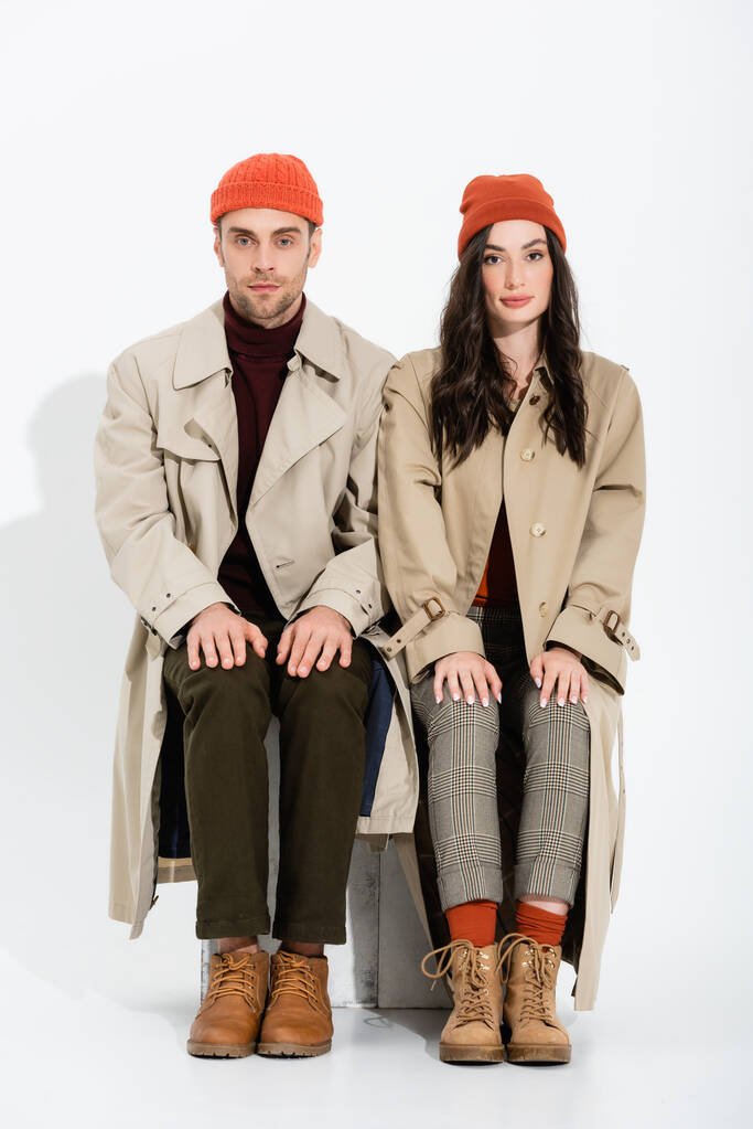 young brunette woman in trench coat sitting near man in beanie hat on white - Photo, Image