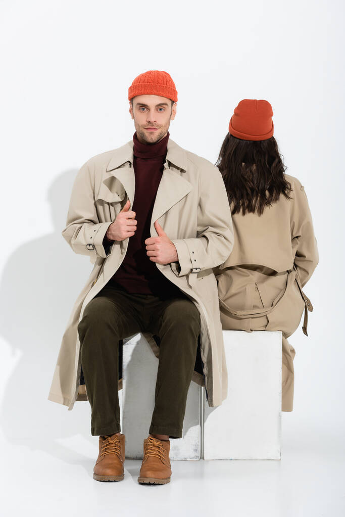 stylischer Mann mit Beanie-Hut berührt Trenchcoat neben Frau, die auf Weiß sitzt - Foto, Bild