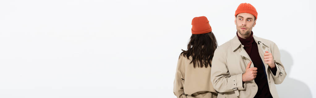 panoramiczny strzał stylowy mężczyzna w czapce dotykając płaszcz okop w pobliżu kobieta izolowane na białym - Zdjęcie, obraz