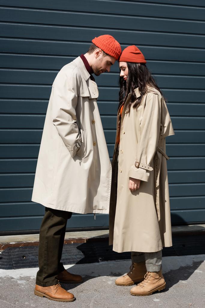 longitud completa de elegante pareja en abrigos de trinchera y sombreros apoyados el uno en el otro fuera - Foto, imagen