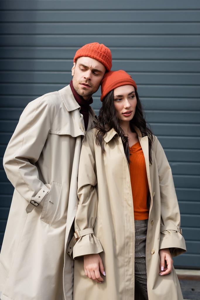 стильний чоловік в траншеї пальто і капелюх стоїть з рукою в кишені біля жінки зовні
 - Фото, зображення
