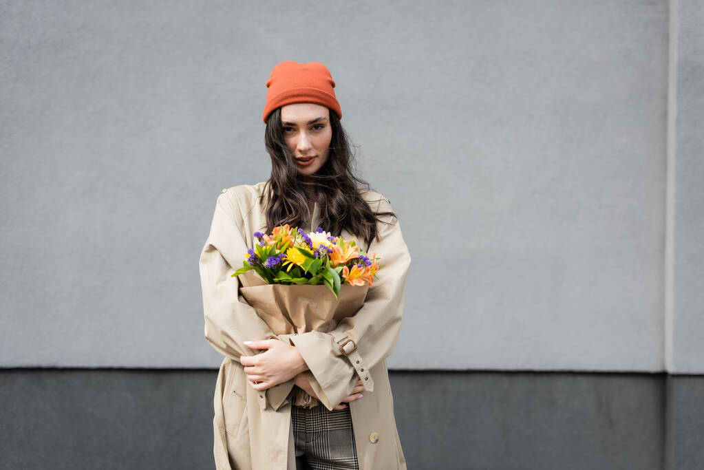 stijlvolle vrouw in muts en trench jas met bloemen in de buurt van grijze muur - Foto, afbeelding