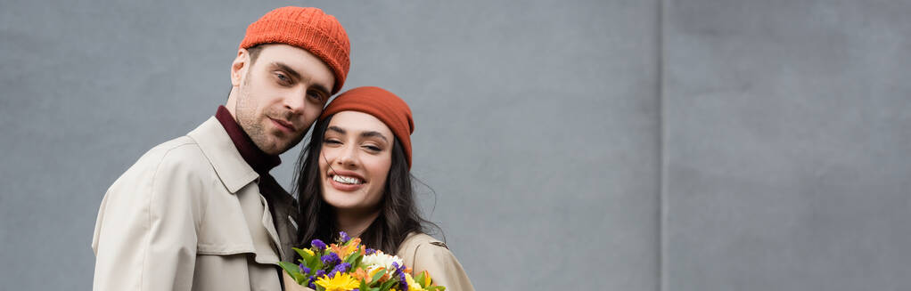 panoramic crop of trendy couple in trench coats and hats near bouquet of flowers  - Photo, Image