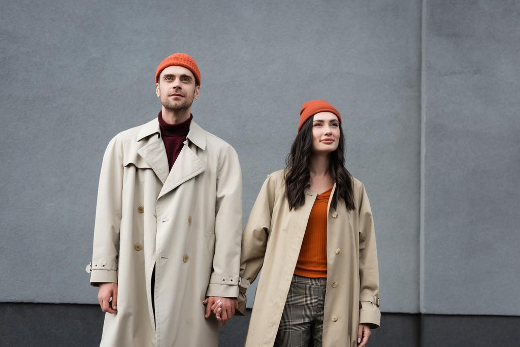 Stilvolles Paar in Trenchcoats und Hüten stehend und Händchen haltend an grauer Wand draußen - Foto, Bild