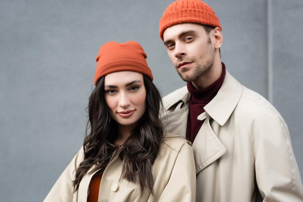 couple à la mode en trench manteaux et chapeaux debout près du mur gris à l'extérieur - Photo, image