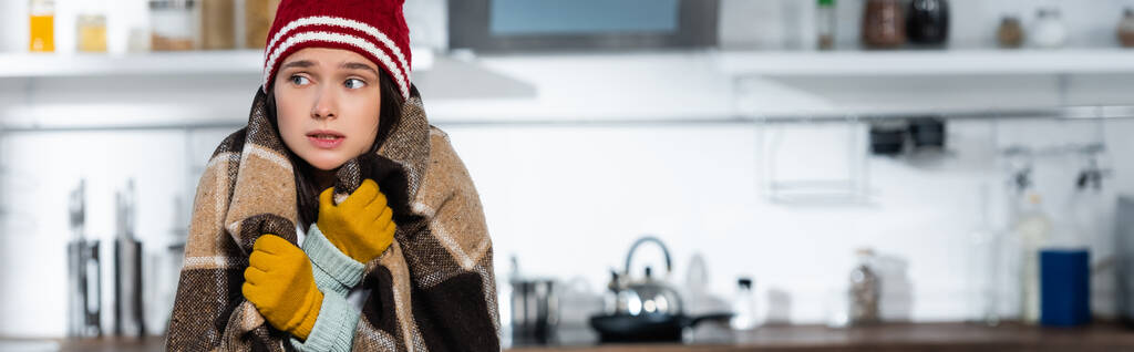 горизонтальное изображение холодной женщины в теплой шляпе и перчатках, покрытой клетчатым одеялом на кухне - Фото, изображение