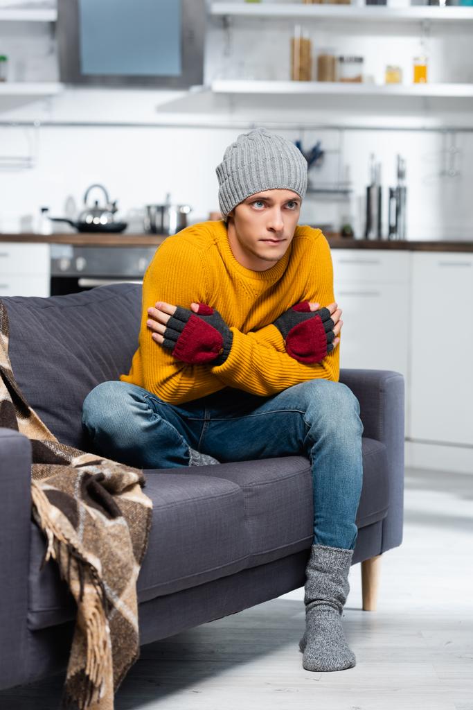 Sıcak şapkalı ve parmaksız eldivenli donan adam soğuk mutfakta kanepede otururken kendine sarılıyor. - Fotoğraf, Görsel