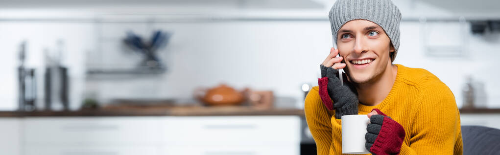 orientation horizontale du jeune homme en bonnet tricoté et gants sans doigts parlant sur smartphone tout en tenant une tasse de boisson chaude à la maison - Photo, image