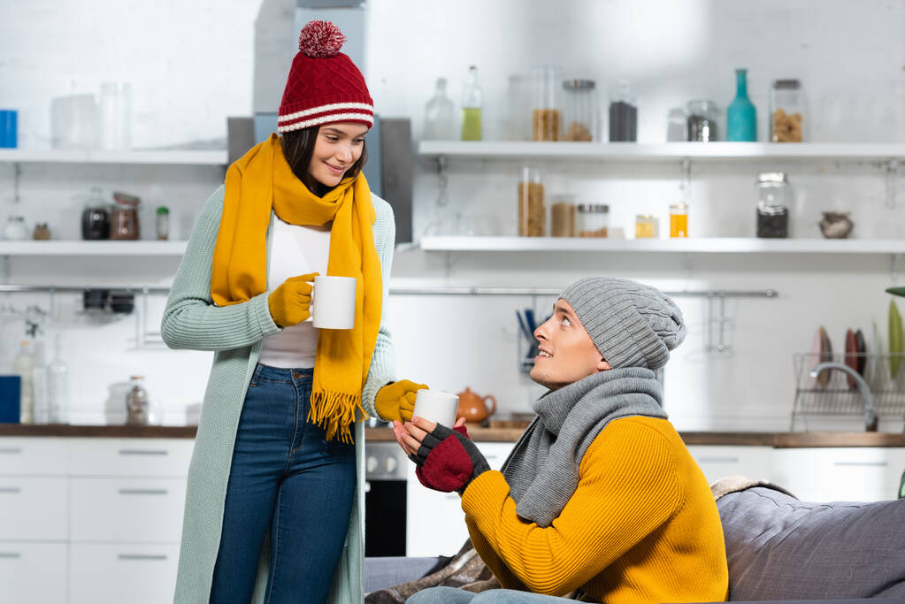 vrouw in gebreide hoed, sjaal en handschoenen geven kopje warme thee aan vriendje zitten op de bank in koude keuken - Foto, afbeelding