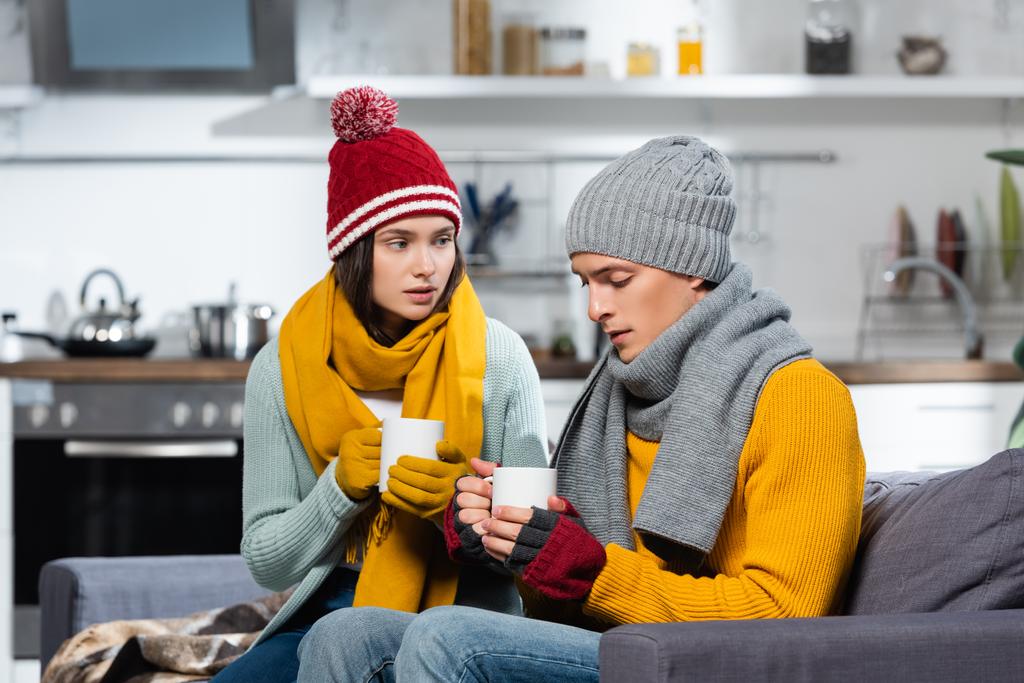 freezing couple in knitted hats, scarfs and gloves holding cups of warming beverage in cold kitchen - Photo, Image