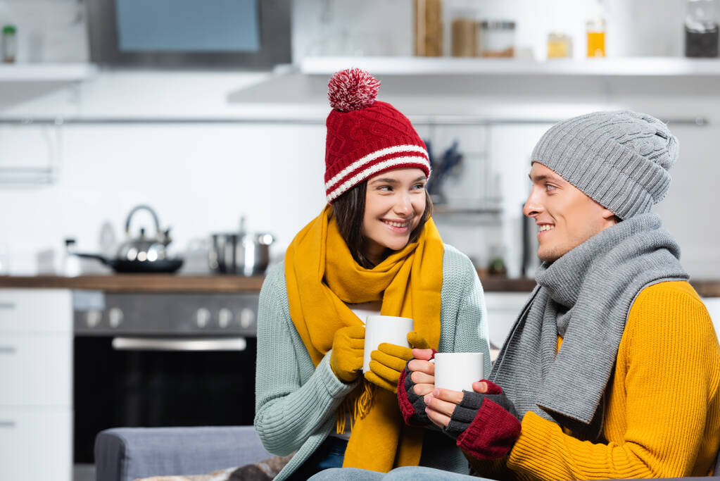 joyful couple in warm hats, scarfs and gloves looking at each other while holding cups of warming beverage in kitchen - Photo, Image