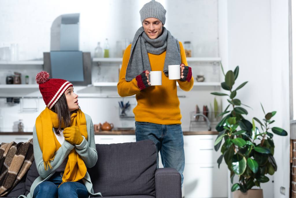 mulher fria em chapéu de malha e luvas olhando para namorado carregando chá quente na cozinha - Foto, Imagem
