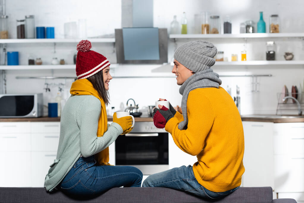 πλαϊνή άποψη του νεαρού ζευγαριού σε ζεστά καπέλα, κασκόλ και γάντια κρατώντας κύπελλα με τσάι στην κουζίνα - Φωτογραφία, εικόνα