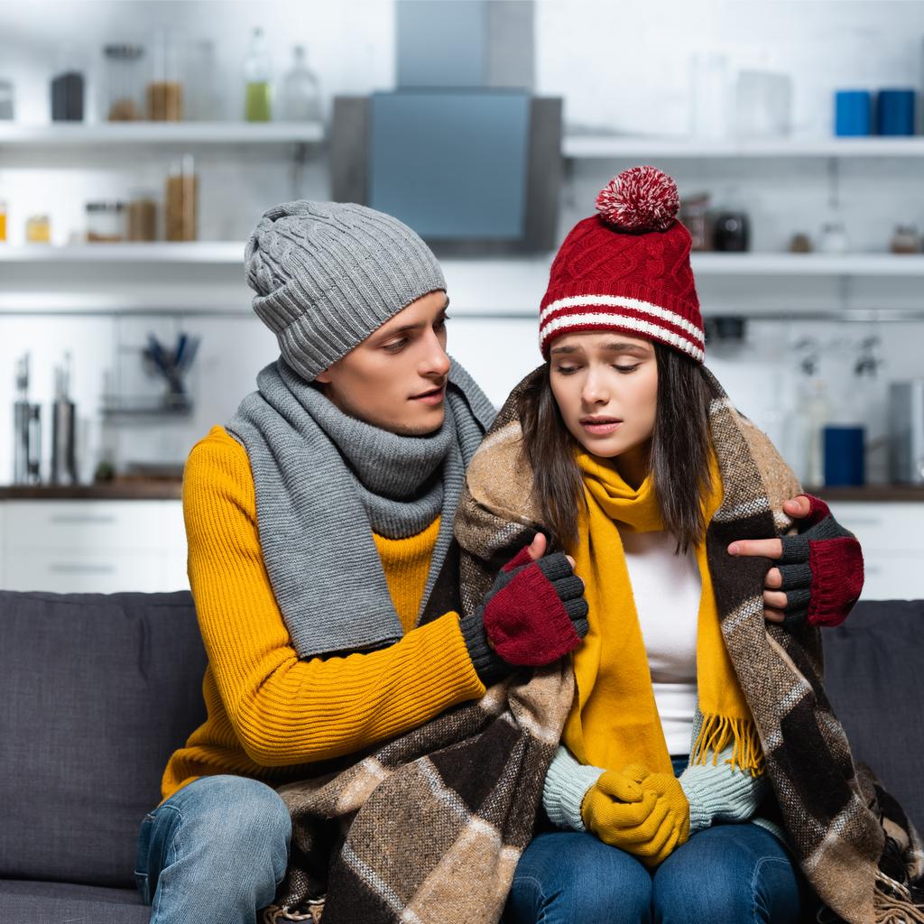 jeune homme en bonnet tricoté et gants couvrant petite amie froide avec couverture à carreaux chaude dans la cuisine - Photo, image