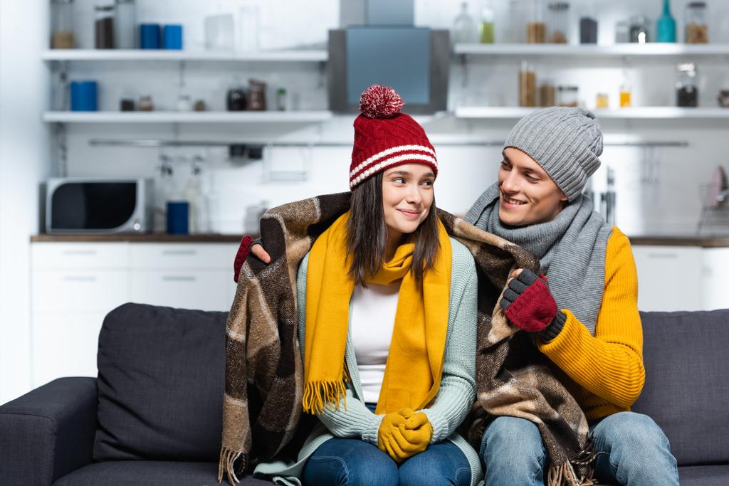 joven en sombrero caliente y guantes que cubren novia congelante con manta a cuadros mientras está sentado en el sofá en la cocina fría - Foto, imagen
