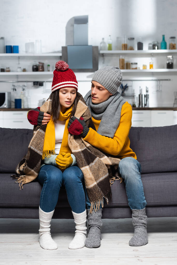 young man in warm hat and gloves covering freezing girlfriend with plaid blanket in cold kitchen - Photo, Image