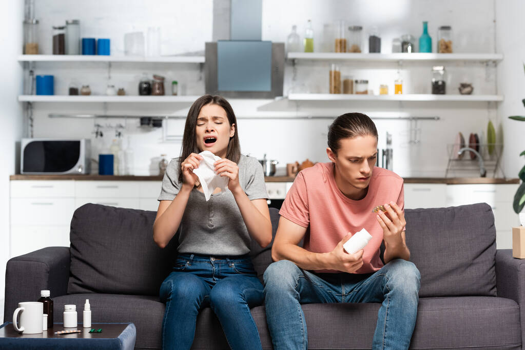chora kobieta kichająca w pobliżu mężczyzny trzymającego butelkę z lekami podczas siedzenia w kuchni - Zdjęcie, obraz