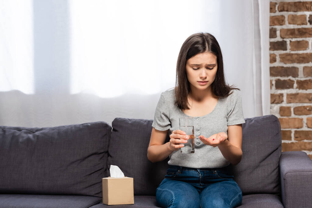 mulher doente segurando vidro de água e pílulas enquanto sentado no sofá perto de guardanapos de papel - Foto, Imagem