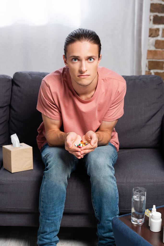 jovem doente segurando um punhado de pílulas enquanto sentado no sofá perto de medicamentos - Foto, Imagem