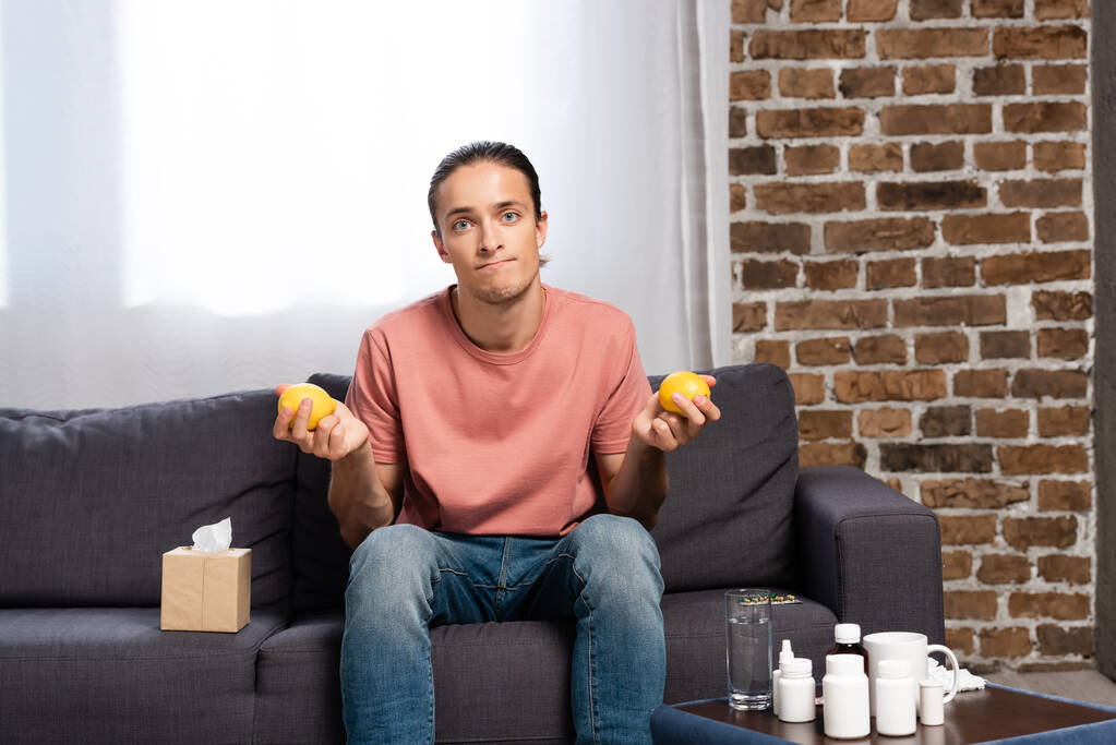 больной человек держит свежие лимоны, сидя на диване возле прикроватного столика с лекарствами - Фото, изображение
