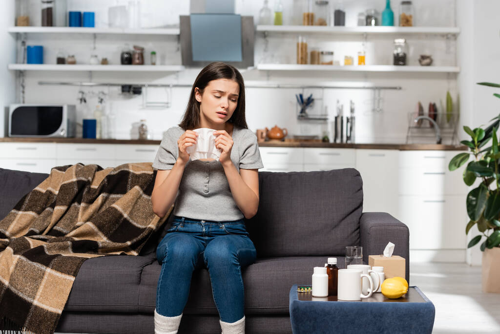 mujer enferma sosteniendo servilleta de papel mientras está sentada en el sofá cerca de manta a cuadros y mesita de noche con medicamentos - Foto, imagen