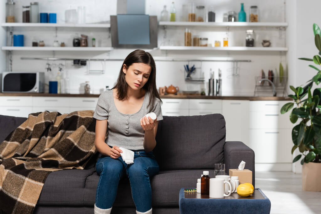donna malata che tiene tovagliolo di carta e bottiglia con pillole mentre siede sul divano vicino al comodino con medicinali - Foto, immagini