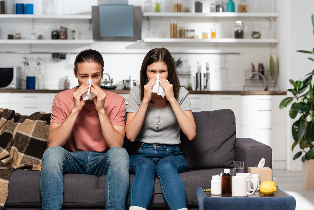 homem doente e mulher espirrando em guardanapos de papel enquanto sentado na cozinha perto de medicamentos - Foto, Imagem