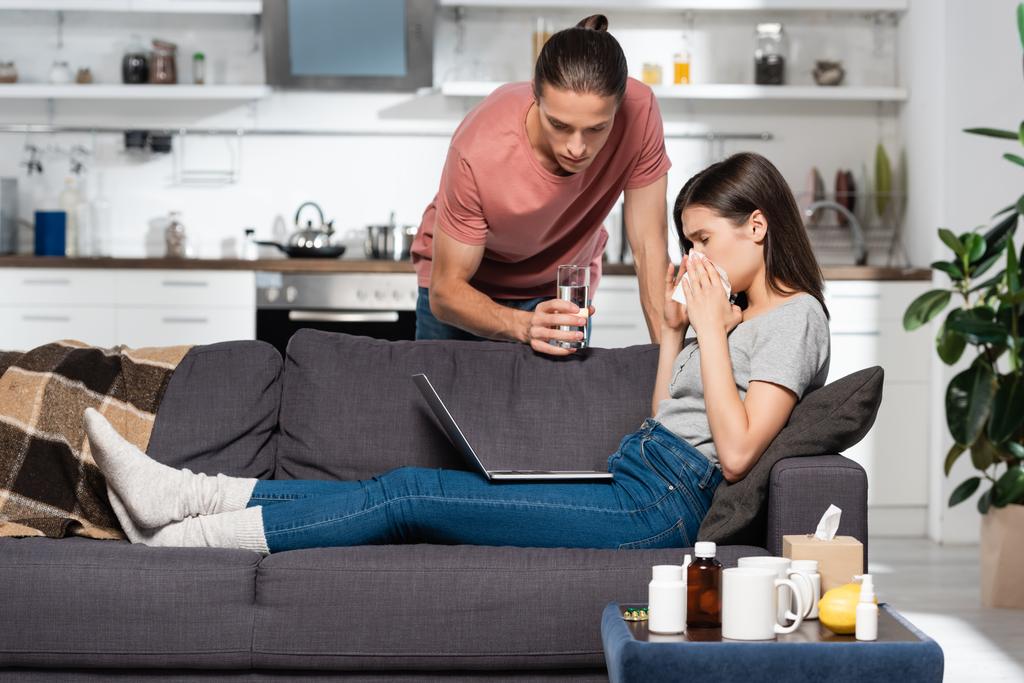 mężczyzna dając szklankę wody chorej kobiecie siedzi na kanapie z laptopem i kicha w papierowej serwetce - Zdjęcie, obraz