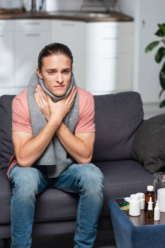 Kranker junger Mann in warmem Schal berührt Halsschmerzen im Sitzen neben Medikamenten - Foto, Bild