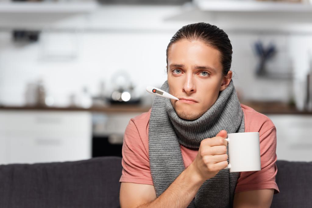 beteg fiatalember meleg sállal a nyakán és hőmérővel a szájában kezében csésze meleg teát, miközben a kamerába néz  - Fotó, kép