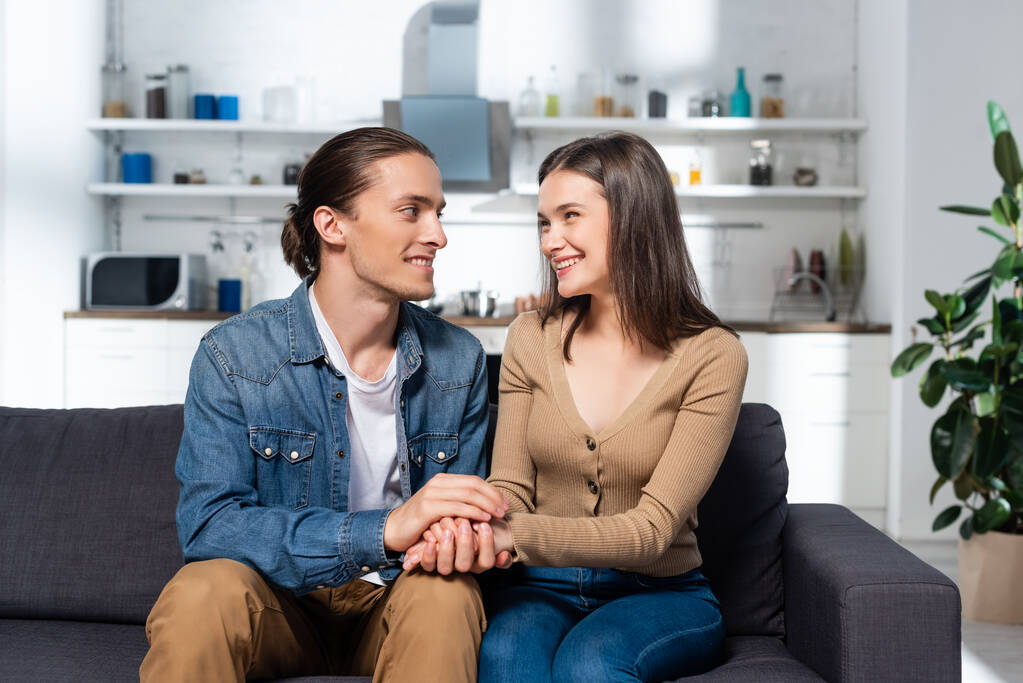 jovem homem tocando mãos de namorada satisfeita enquanto sentado no sofá na cozinha juntos - Foto, Imagem