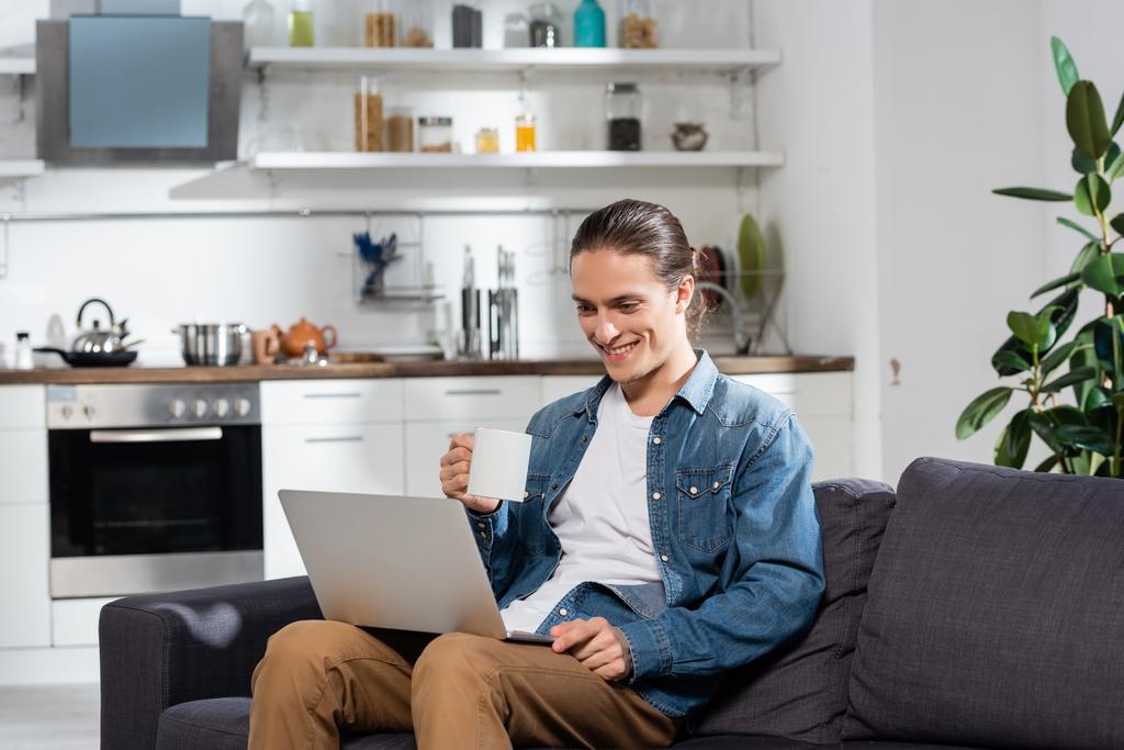 young freelancer holding cup of tea while sitting on sofa in kitchen and using laptop - Photo, Image
