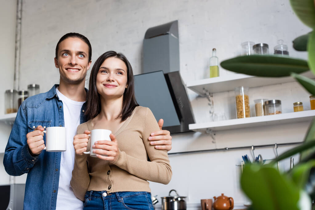 επιλεκτική εστίαση χαρούμενη ζευγάρι κοιτάζοντας μακριά, ενώ κρατώντας φλιτζάνια του καφέ στην κουζίνα - Φωτογραφία, εικόνα