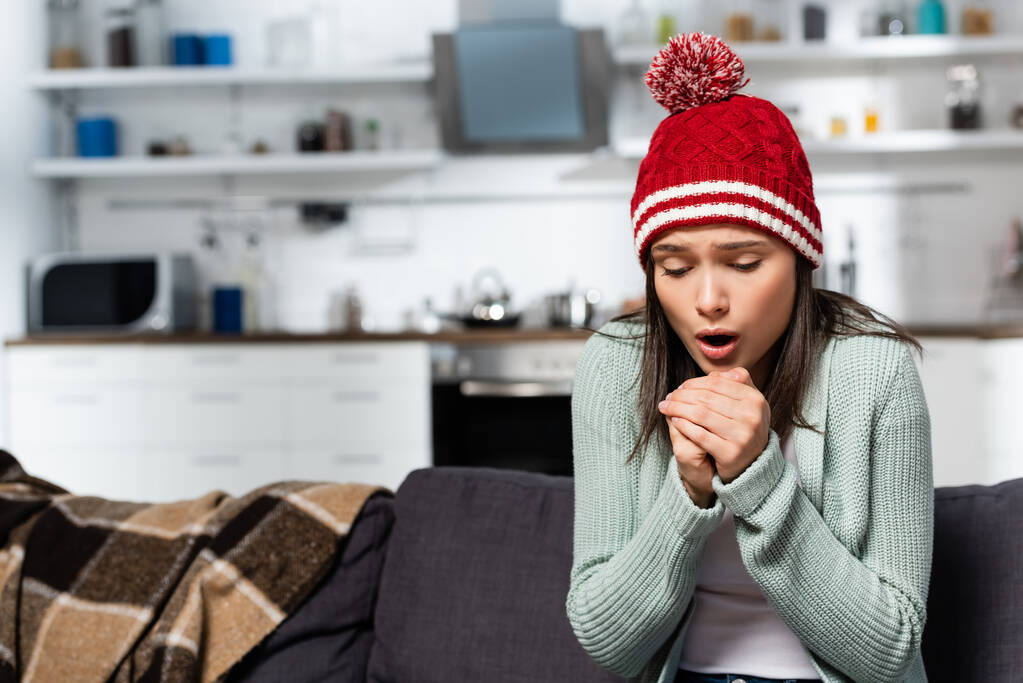κρύα γυναίκα σε ζεστό καπέλο φυσάει σε σφιγμένα χέρια, ενώ κάθεται στον καναπέ στην κουζίνα - Φωτογραφία, εικόνα