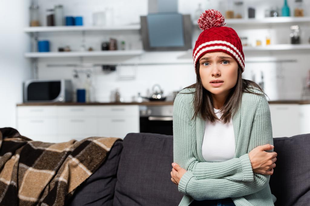 freezing woman in knitted hat hugging herself while sitting in cold kitchen - Photo, Image