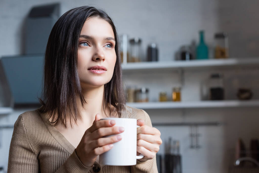 álmodozó barna nő néz félre, miközben tartja csésze teát otthon - Fotó, kép