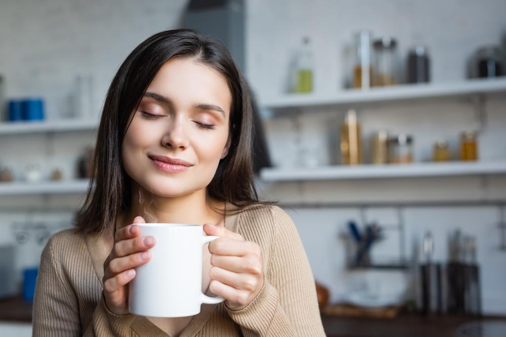 zadowolona młoda kobieta z zamkniętymi oczami trzyma filiżankę ciepłej herbaty w domu - Zdjęcie, obraz