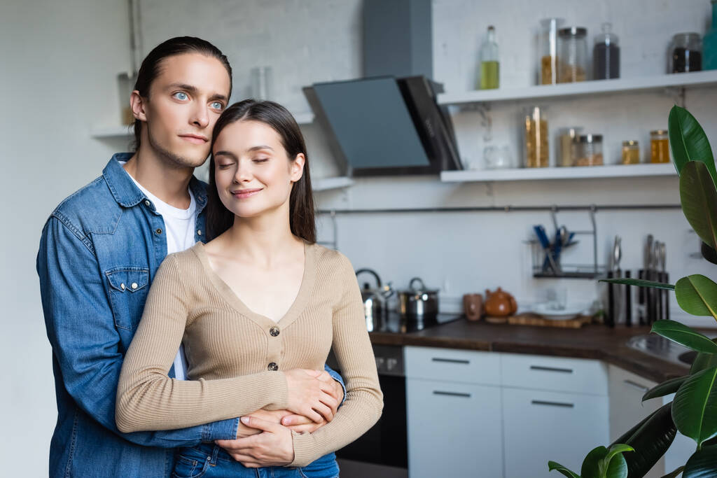 νεαρός άνδρας αγκαλιάζει γυναίκα στέκεται στην κουζίνα με κλειστά μάτια - Φωτογραφία, εικόνα