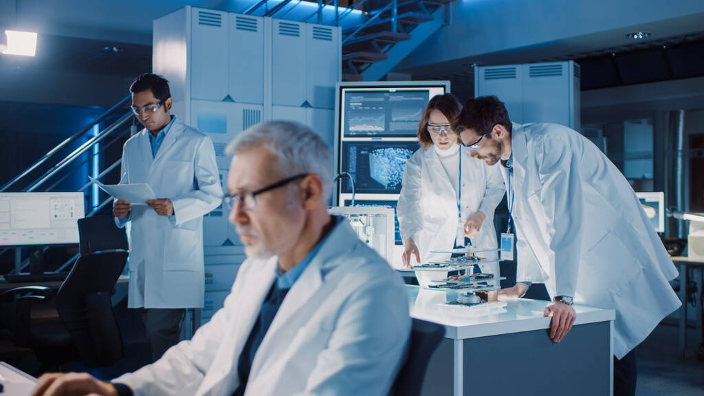 Διαφορετική διεθνής ομάδα βιομηχανικών επιστημόνων και μηχανικών που φορούν λευκά παλτά και εργάζονται στο σχεδιασμό βαρέων μηχανημάτων στο ερευνητικό εργαστήριο. Επαγγελματίες που χρησιμοποιούν 3D εκτυπωτή, υπολογιστές και μικροσκόπιο - Φωτογραφία, εικόνα